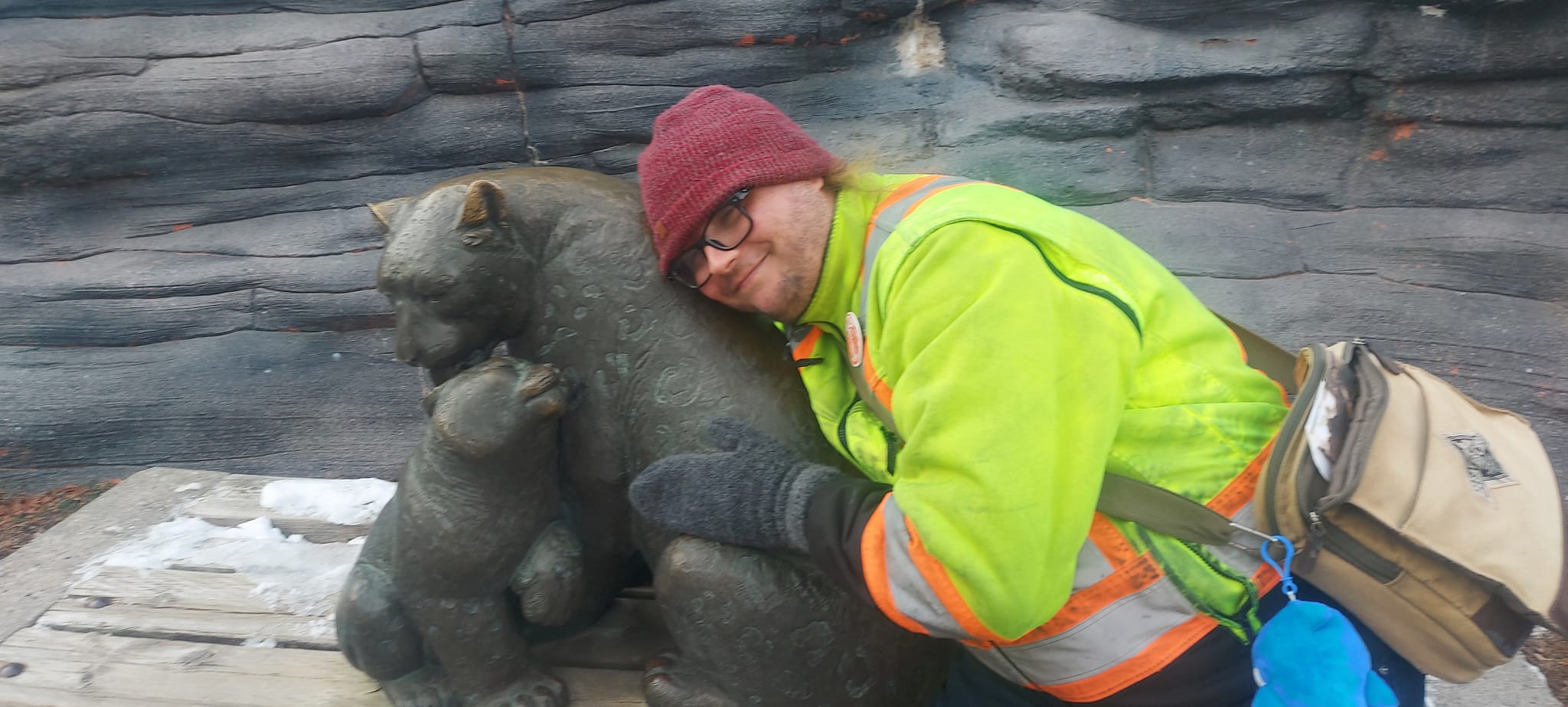 A man wearing a red toque and a high visibility winter coat sits next to a snow leopard and kitten statue, embracing the mother cat